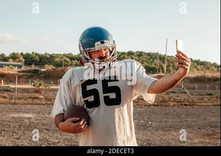 Joueur américain de football utilisant un smartphone pour prendre un selfie tout en tenant un ballon à l'extérieur. Banque D'Images
