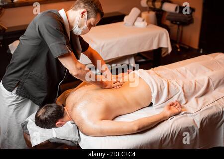 Masseur professionnel en masque facial faisant un massage profond à un client masculin au salon Spa. Les affaires pendant le concept de l'épidémie Banque D'Images
