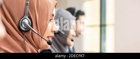 Femme musulmane asiatique portant un micro-casque qui travaille comme service à la clientèle opérateur avec équipe dans l'arrière-plan de la bannière du bureau du centre d'appels Banque D'Images