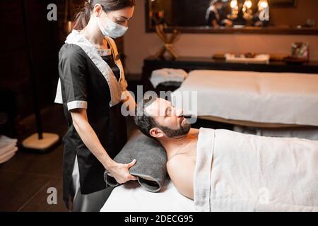 Masseuse professionnelle féminine en masque médical faisant un massage facial à un client masculin au salon de Spa. Les affaires pendant le concept de l'épidémie Banque D'Images