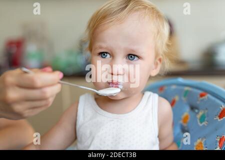 Mère nourrissant un adorable petit garçon blond caucasien avec du yaourt ou du fromage cottage au lait pour le déjeuner. Enfant mangeant en chaise haute à Banque D'Images