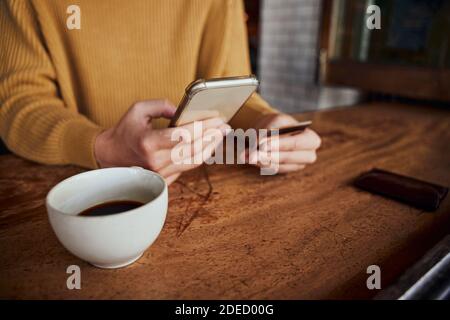 Gros plan de jeunes femmes mains tenant un smartphone et une carte bancaire tout en effectuant le paiement en ligne dans le café boire du café Banque D'Images