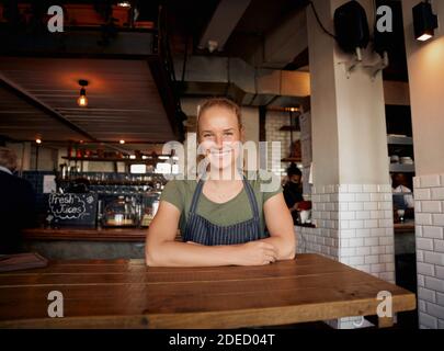Jeune femme gaie propriétaire de café portant un tablier souriant regardant et de se pencher sur la table Banque D'Images