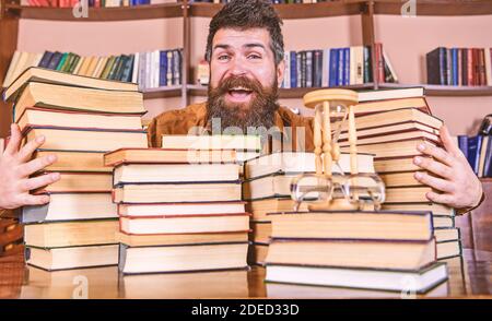 Homme sur le visage heureux entre des piles de livres, tout en étudiant dans la bibliothèque, les étagères sur le fond. Concept de bibliothécaire. Professeur ou étudiant avec la barbe assis à table avec des livres, défoqué. Banque D'Images