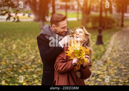 Des jeunes heureux amoureux, un homme embrassant une femme de derrière elle en traquant la joue, un couple heureux marchant dans un parc d'automne portant des manteaux élégants et la cueillette Banque D'Images