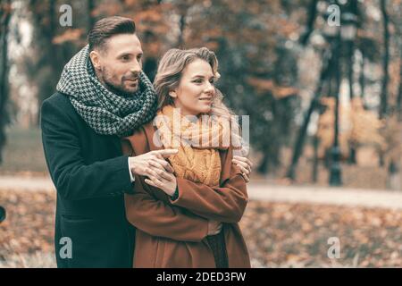 Homme embrassant femme debout derrière elle regardant une direction latéralement dans le parc d'automne. Photo en plein air d'un jeune couple amoureux qui a passé un excellent moment Banque D'Images