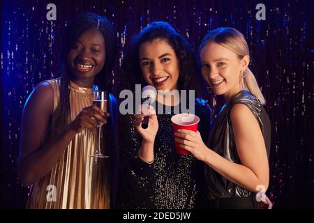 Portrait de trois jeunes femmes tenant des verres de champagne et souriant à l'appareil photo tout en posant sur un fond étincelant lors d'une soirée karaoké Banque D'Images