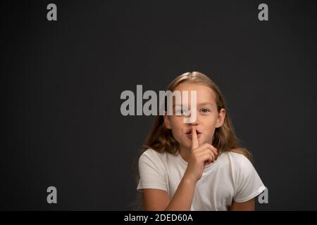 Gardez le calme 8,10 ans fille de tenir son doigt sur sa bouche disant à tout le monde de garder le silence en portant le t blanc maillot souriant à l'appareil photo isolé Banque D'Images