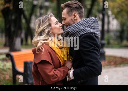 Un jeune homme de couple tient ses amies les mains et les nez en contact se tiennent dans le parc de ville d'automne portant des manteaux et des foulards. Couple souriant qui se serre Banque D'Images