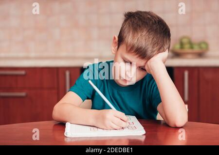 7 yeas vieux garçon assis à table dans la cuisine et écrivant des notes sur les feuilles, les devoirs difficiles, l'enfant se concentrant sur des exemples Banque D'Images