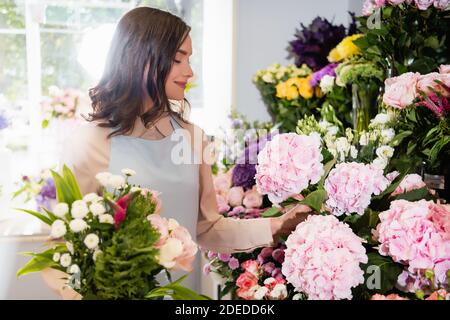 Fleuriste souriant avec bouquet prenant hortensia dans fleuriste avec fenêtre floue sur l'arrière-plan Banque D'Images