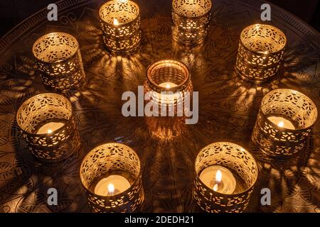 Neuf bougies décoratives éclairées, 8 petits chandeliers en métal entourant un chandelier en verre au milieu d'une table ronde en métal, l'éclairage se reflète Banque D'Images