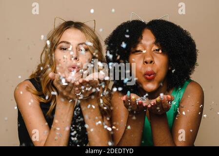 Les jeunes femmes noires et caucasiennes soufflent des confettis dans l'appareil photo sur un fond beige. Fêtez Noël en studio. Concept de fête, d'événement et de célébration. Banque D'Images