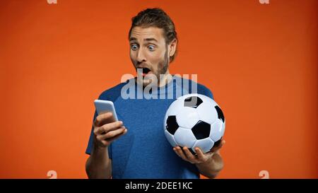 fan de football choqué dans un t-shirt bleu avec smartphone et tenue ballon de football sur orange Banque D'Images