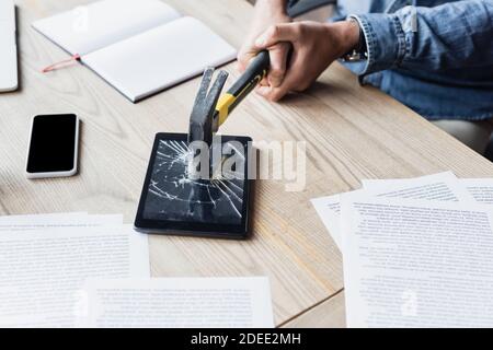 Vue rognée d'un homme d'affaires frappant une tablette numérique avec un marteau pendant assis sur le lieu de travail avec des documents Banque D'Images
