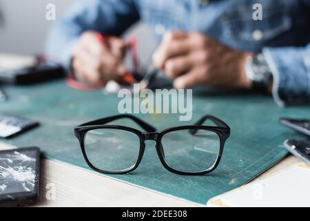 Vue rapprochée des lunettes avec cadre noir sur la table près de téléphones mobiles cassés avec un réparateur flou sur l'arrière-plan Banque D'Images