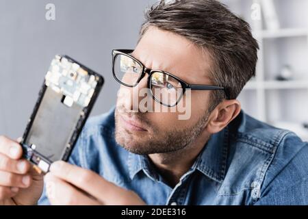 Réparateur concentré dans les lunettes regardant la partie démontée du mobile téléphone au premier plan flou Banque D'Images