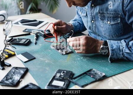 Vue rognée du réparateur qui maintient les capteurs du multimètre sur le démontable partie du téléphone portable sur la table Banque D'Images