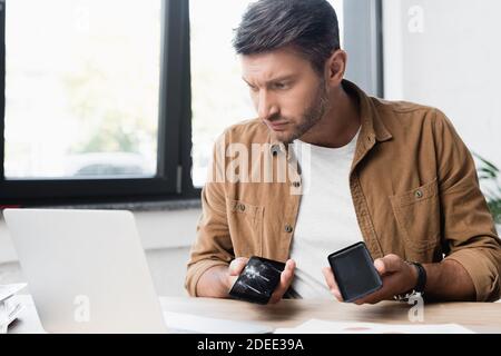 Homme d'affaires sérieux avec un smartphone cassé démonté, regardant l'ordinateur portable sur le lieu de travail sur un arrière-plan flou Banque D'Images