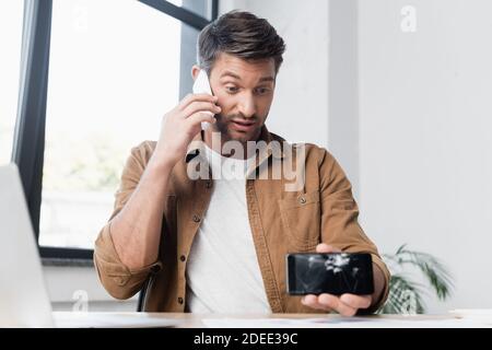 Homme d'affaires avec un smartphone écrasé parlant sur un téléphone mobile pendant qu'il est assis sur le lieu de travail, sur un arrière-plan flou Banque D'Images