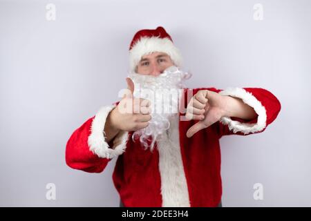 Homme habillé comme le Père Noël debout sur un fond blanc isolé faisant des pouces de haut en bas, désaccord et expression d'accord. Un conflit fou Banque D'Images