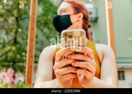 Jeune femme assise à l'extérieur, envoyant des SMS au téléphone, portant un masque facial. Banque D'Images