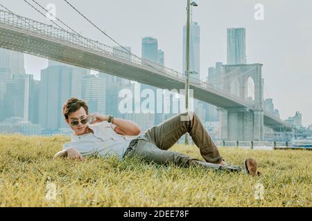 Jeune homme assis à l'extérieur dans le parc portant des lunettes de soleil. Banque D'Images