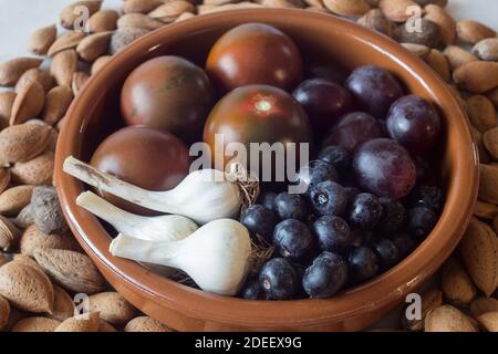 Gros plan d'un bol en argile rond fait à la main rempli de produits biologiques, de têtes d'ail, de bleuets, de raisins et de tomates entourés d'amandes entières. Banque D'Images