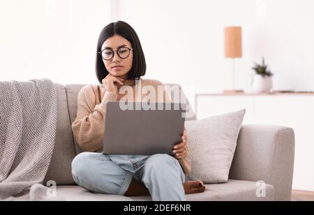 Femme asiatique assise sur un canapé, utilisant un ordinateur portable et pensant Banque D'Images
