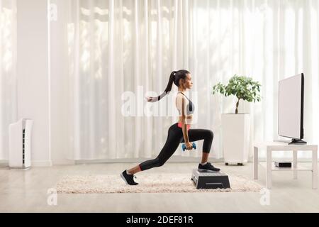 Jeune femme faisant de l'exercice STEP aérobie avec de petits poids à l'avant d'une télévision dans un salon Banque D'Images