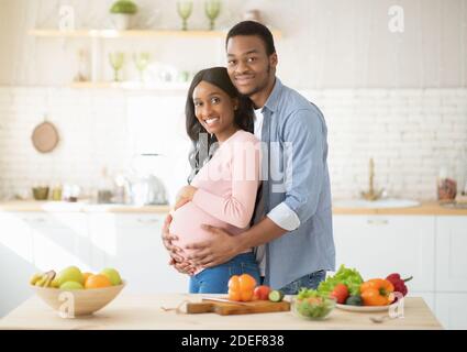 Régime alimentaire pour une grossesse en santé. Homme noir aimant embrassant sa magnifique petite amie expectative en cuisinant dans la cuisine Banque D'Images