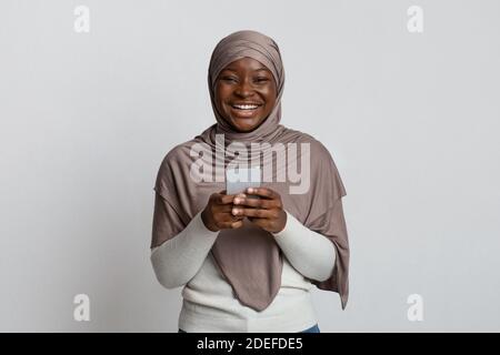 Joyeuse Black Muslim Lady en hijab tenant le smartphone et en riant À la caméra Banque D'Images