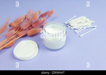 Crème hydratante en pot en verre ouvert, herbes roses Lagurus, tampons de coton et bâtons sur fond violet clair texturé. Vue avant. Bio bio naturel e Banque D'Images