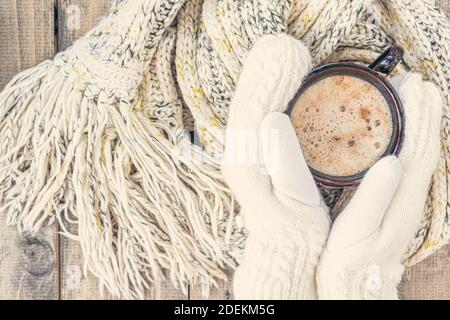 Les mains des femmes en moufles blanches tiennent une tasse de café chaud avec mousse sur une table en bois avec un foulard en laine à maille chaude. Arrière-plan de Noël.concept d'hiver, wa Banque D'Images