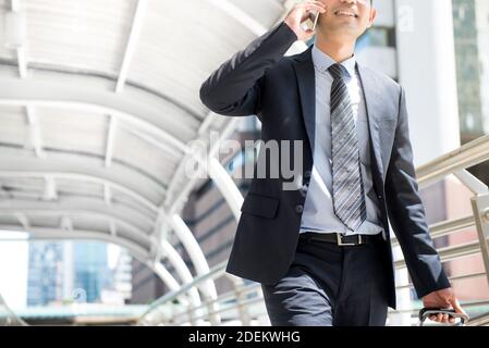 Homme d'affaires qui appelle sur un téléphone portable tout en tirant ses bagages et en marchant dans un passage couvert extérieur - affaires sur la route et concept de déplacement Banque D'Images