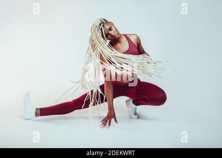 Femme afro-américaine avec de longs cheveux tressés faisant des jambes s'étire sur le sol. Athlète féminine s'exerçant sur des exercices de flexibilité. Banque D'Images