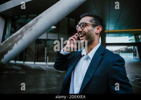 Homme d'affaires de course mixte habillé chatter intelligemment sur un appareil mobile riant tout en marchant vers le travail Banque D'Images