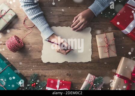 Femme dans un chandail gris écrivant sur papier d'artisanat avec un crayon rouge au centre des cadeaux de noël une table en bois avec du pin et du gui Banque D'Images