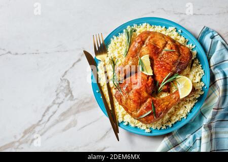 Des quartiers de cuisses de poulet servis sur le millet sur une assiette avec des couverts dorés, des quartiers de citron et des brins de romarin frais sur un fond de pierre en marbre blanc Banque D'Images