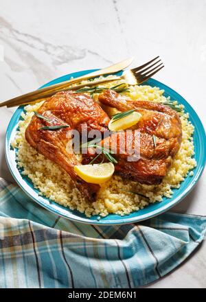 Des quartiers de cuisses de poulet servis sur le millet sur une assiette avec des couverts dorés, des quartiers de citron et des brins de romarin frais sur un fond de pierre en marbre blanc Banque D'Images