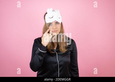 Jeune femme caucasienne portant un masque de sommeil et un pyjama sur fond rose isolé pointant vers l'œil vous regardant le geste, expression suspecte Banque D'Images