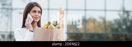 femme souriante parlant sur un téléphone portable tout en tenant le sac de shopping avec de la nourriture à l'extérieur, bannière Banque D'Images