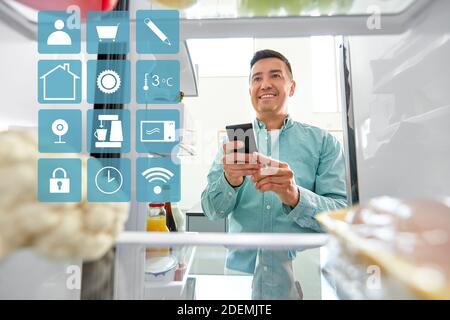 L'homme avec le smartphone faisant liste des aliments dans un réfrigérateur Banque D'Images