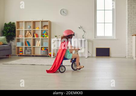 Jeune fille d'âge préscolaire dans un casque et cape rouge à cheval sur son tricycle dans une chambre confortable pour bébé Banque D'Images