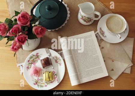 CORK, IRLANDE - 18 novembre 2020 : café et mini-gâteaux servis sur un thé d'époque avec roses roses roses, théière en fer et un livre à lire. Banque D'Images