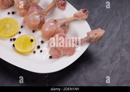 Lollipop de poulet cru, six morceaux de Lollipop de poulet disposés sur un servir une assiette blanche et garni de tranches de citron et de noir poivre sur texture grise Banque D'Images