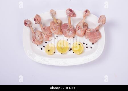 Lipop de poulet cru, six morceaux de lipop de poulet disposés sur une assiette blanche de service avec des tranches de citron et du poivre noir sur fond blanc Banque D'Images