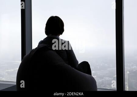 Silhouette d'une femme assise sur un sac de beanbag devant une fenêtre panoramique et vue sur la ville brumeuse Banque D'Images