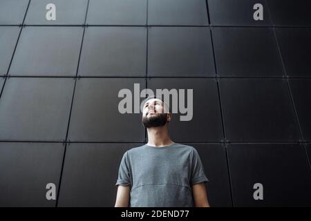 Portrait d'un beau gars élégant regardant vers le haut, taille basse portant un t-shirt vide gris debout sur fond de mur noir. Banque D'Images