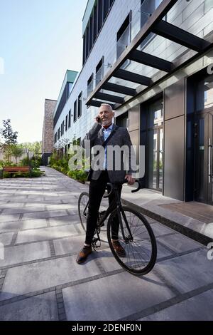 Photo en longueur d'un homme d'affaires mûr souriant tout en parlant au téléphone, debout avec son vélo près d'un immeuble de bureau moderne à l'extérieur Banque D'Images